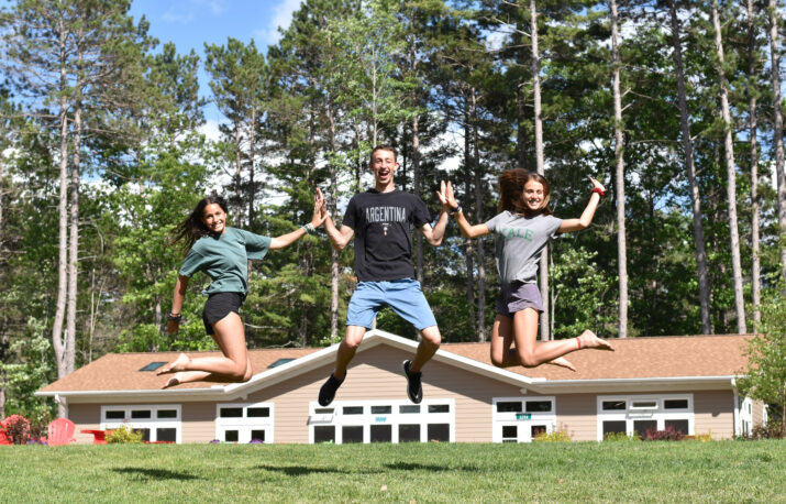 Two girls and a boy in the middle jump in the air in front of a cabin and lots of tall trees.