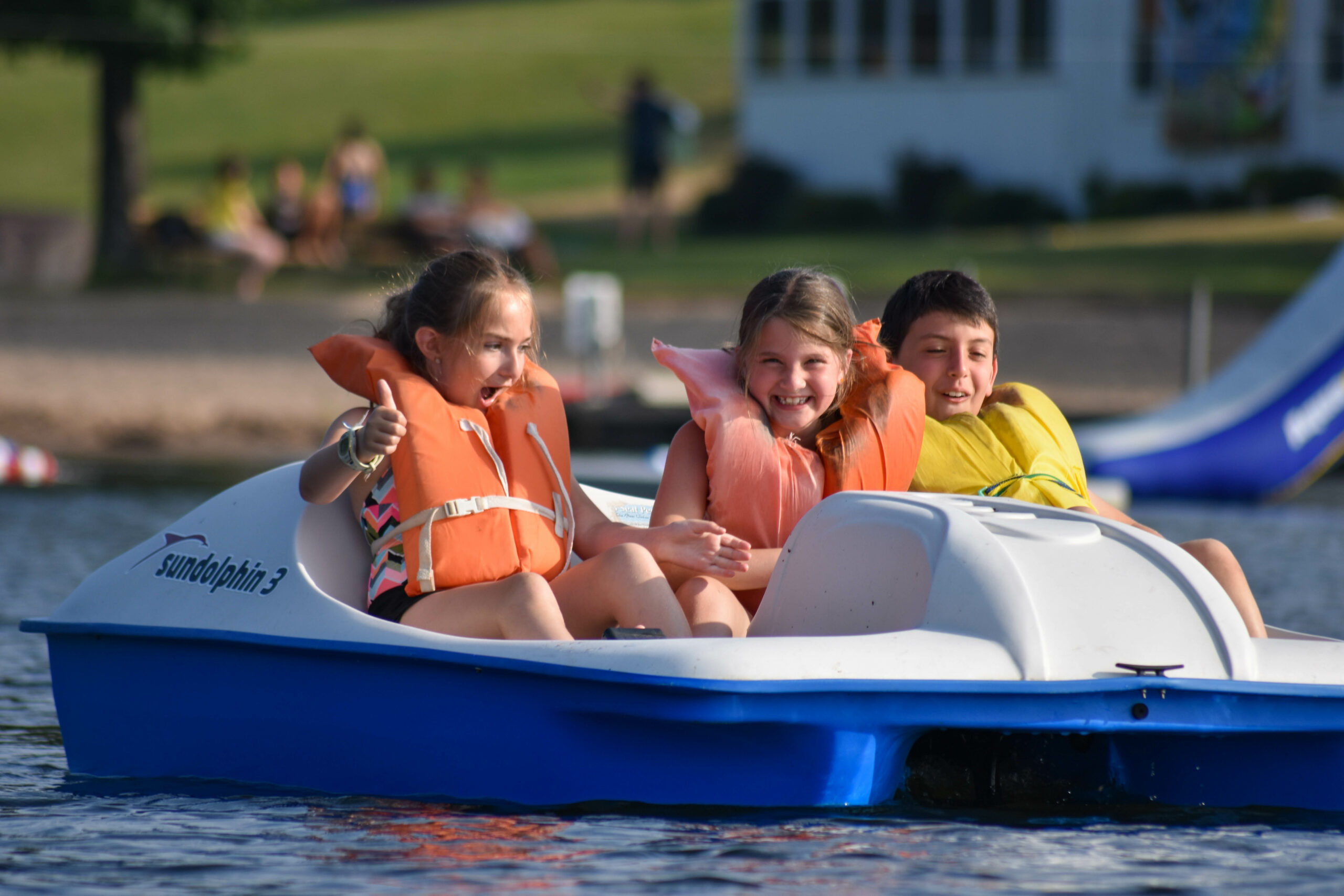 Two young girls and a boy in orange life jackets paddle a paddle boat.